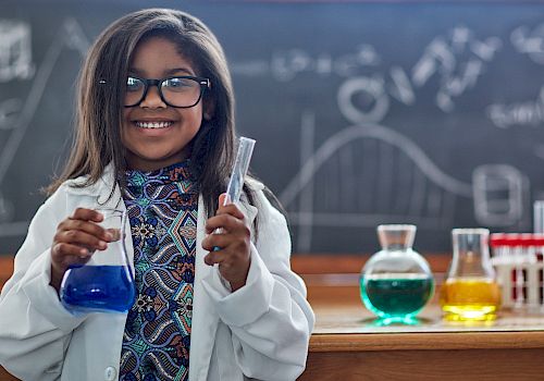 A girl in a lab coat holds science equipment in a classroom with colorful liquids and a chalkboard in the background, smiling.