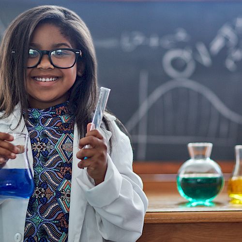 A girl in a lab coat holds science equipment in a classroom with colorful liquids and a chalkboard in the background, smiling.