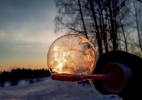 The image shows a frozen bubble on a wand, with a snowy landscape and trees in the background, illuminated by a sunset.