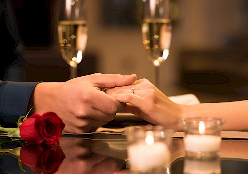 A couple holding hands over a table, with a rose, two candles, and champagne glasses in the background, creating a romantic setting.