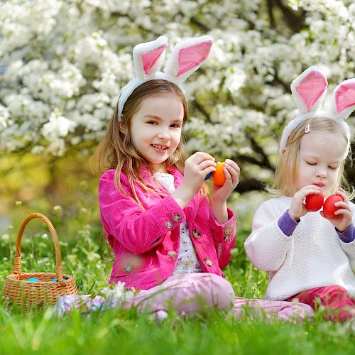 Two children with bunny ears sit on grass holding Easter eggs near a basket, with blooming trees in the background.
