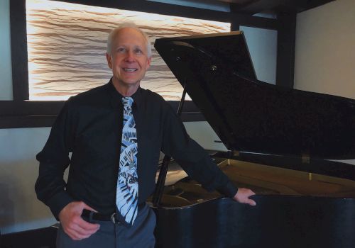 A man in a black shirt and patterned tie stands next to a grand piano in a modern room with abstract wall art, smiling at the camera.