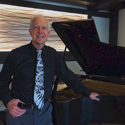 A man in a black shirt and patterned tie stands next to a grand piano in a modern room with abstract wall art, smiling at the camera.
