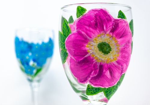 Two wine glasses with painted floral designs; one has a large pink flower, and the other features blue flowers in the background, ending the sentence.