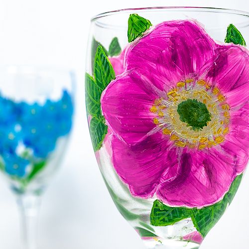 Two wine glasses with painted floral designs; one has a large pink flower, and the other features blue flowers in the background, ending the sentence.