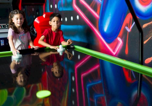 Two kids are playing air hockey surrounded by colorful neon lights and graffiti in an arcade setting.