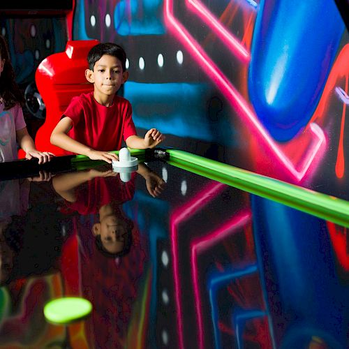 Two kids are playing air hockey surrounded by colorful neon lights and graffiti in an arcade setting.
