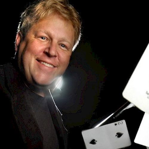 A person is smiling while holding playing cards, including the ace of spades, in a dynamic pose against a dark background.