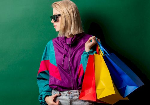 A person wearing sunglasses and a colorful jacket holds several bright shopping bags, standing against a green background.