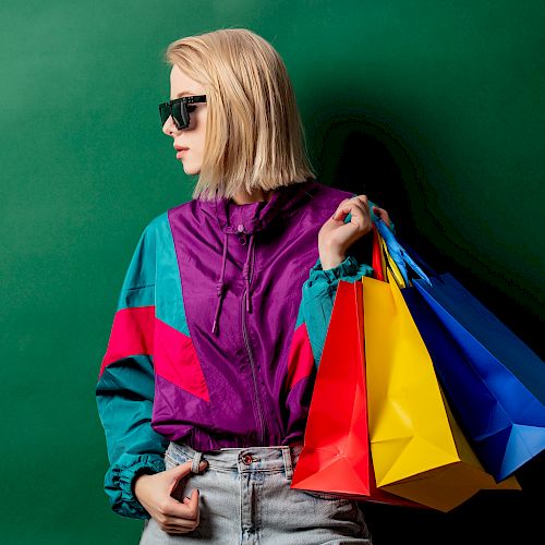 A person wearing sunglasses and a colorful jacket holds several bright shopping bags, standing against a green background.