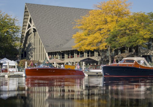 The image shows boats docked in a serene lake with a large A-frame building and autumnal trees in the background, reflecting on the water’s surface.