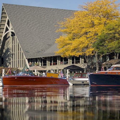 The image shows boats docked in a serene lake with a large A-frame building and autumnal trees in the background, reflecting on the water’s surface.