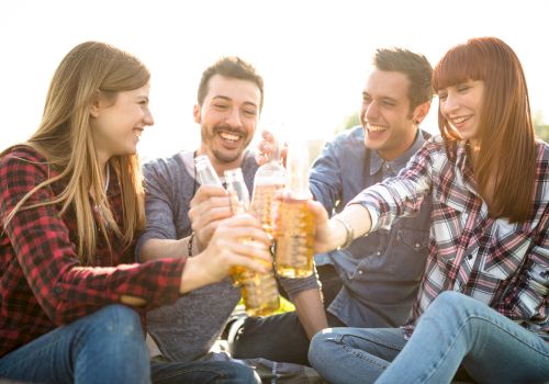Four friends are sitting outdoors, smiling and raising their drinks in a toast. They appear to be enjoying a casual and joyful moment together.