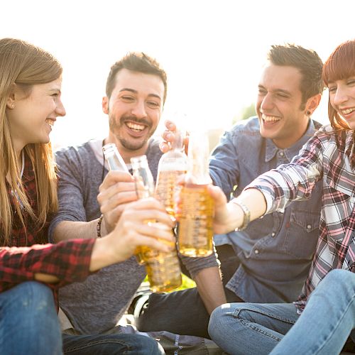 Four friends are sitting outdoors, smiling and raising their drinks in a toast. They appear to be enjoying a casual and joyful moment together.