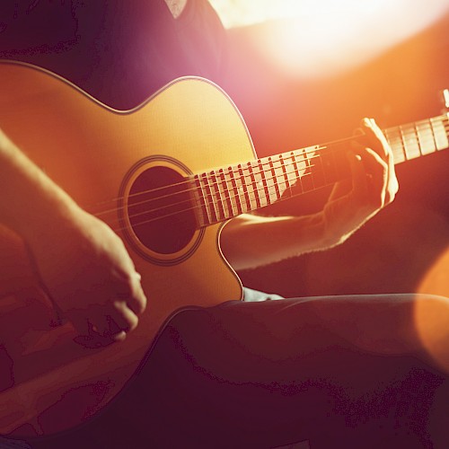 A person is playing an acoustic guitar in a warmly lit setting, with a focus on their hands on the strings.