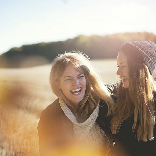 Two women are outdoors in a field, smiling and laughing with their arms around each other, enjoying a sunny day in nature.