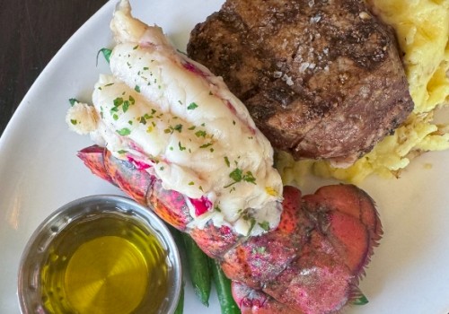 The image shows a plated meal with a steak, lobster tail, mashed potatoes, green beans, and a small dish of melted butter for dipping.