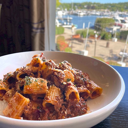 A bowl of rigatoni pasta with meat sauce sits on a table, with a scenic marina and boats visible through the window in the background.