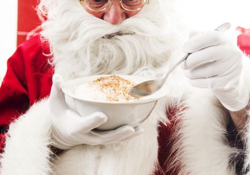 An individual dressed as Santa Claus is holding a bowl and eating with a spoon, appearing to be enjoying a meal, likely porridge or soup.