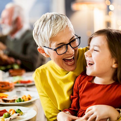 An older woman with glasses and short white hair laughs with a young girl in a red dress at a festive table. They appear to be enjoying a meal together.