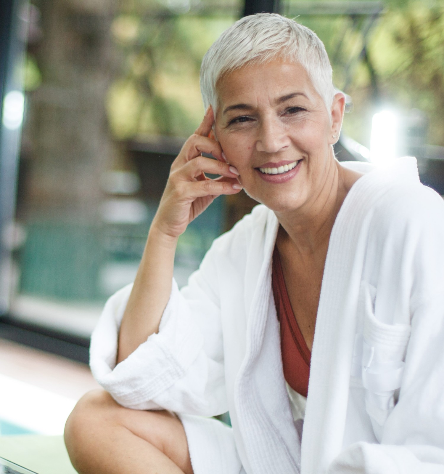 A person with short hair is smiling, wearing a white robe, sitting by a poolside, with blurred trees visible through glass windows.