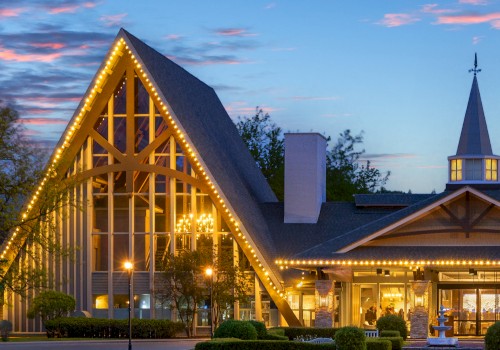 A building with a tall, pointed roof and lit with string lights stands against a sunset sky, featuring a steeple and large windows.