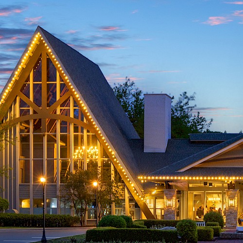 A building with a tall, pointed roof and lit with string lights stands against a sunset sky, featuring a steeple and large windows.