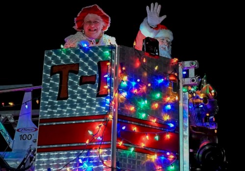 Santa and Mrs. Claus are smiling and waving from a fire truck decorated with colorful lights at night.