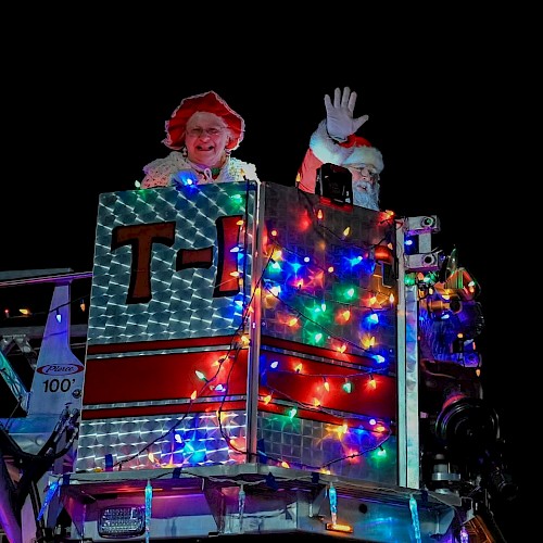 Santa and Mrs. Claus are smiling and waving from a fire truck decorated with colorful lights at night.