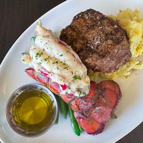 The image shows a plated meal with a steak, lobster tail, mashed potatoes, green beans, and a small dish of melted butter for dipping.