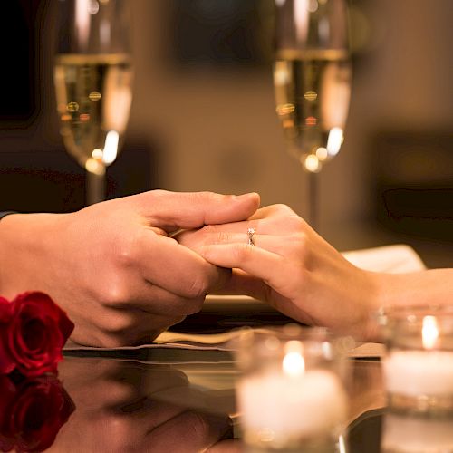 A couple holding hands over a table, with a rose, two candles, and champagne glasses in the background, creating a romantic setting.