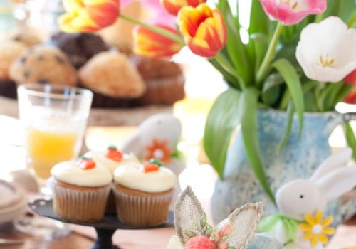 A colorful table setting with tulips, cupcakes, juice, and a rabbit figurine, featuring a napkin with a butterfly decor detail.