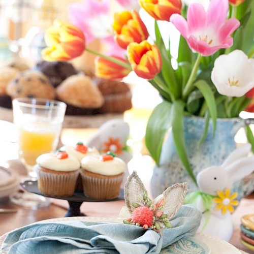 A colorful table setting with tulips, cupcakes, juice, and a rabbit figurine, featuring a napkin with a butterfly decor detail.