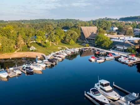 Aerial view of a marina with several boats docked, surrounded by lush greenery and buildings. Calm waters reflect the serene environment.