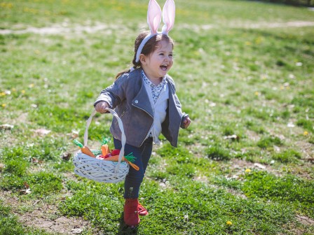 A child is happily running outdoors, wearing bunny ears and holding a basket with decorative items, likely celebrating Easter or a similar event.
