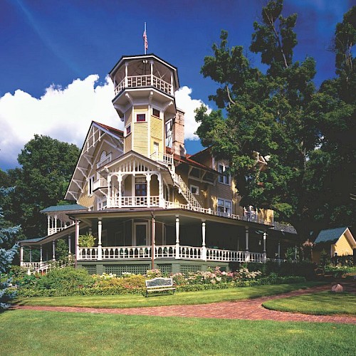 Victorian-style house with a tall tower, surrounded by greenery and a clear blue sky, featuring a charming wraparound porch.