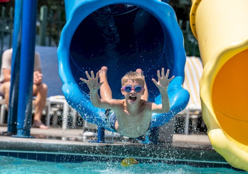 A child splashes out of a blue water slide into a pool, smiling and wearing goggles, with another yellow slide beside it.