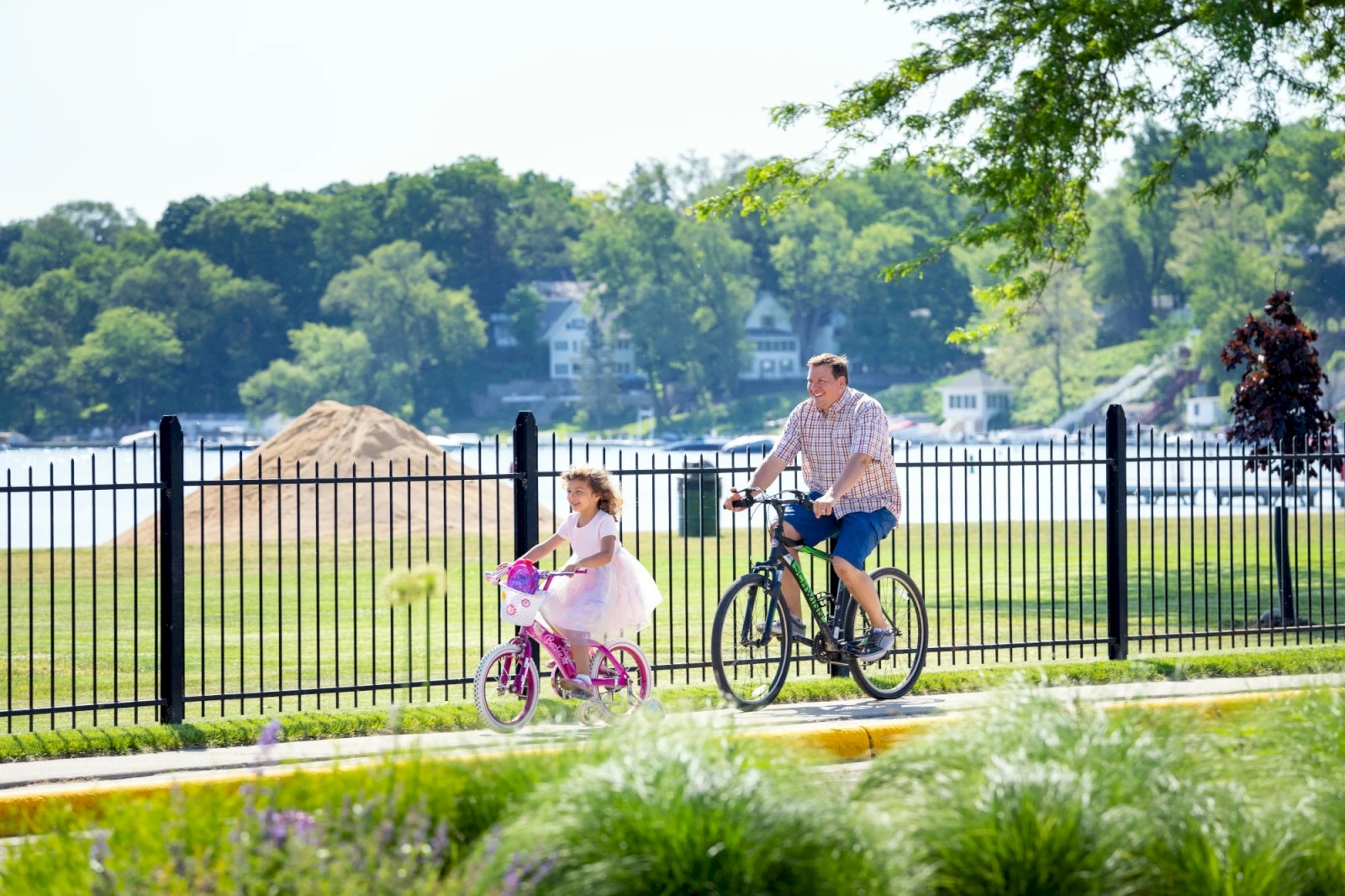 A child and an adult are riding bikes on a path near a fence, with a scenic lake and greenery in the background.