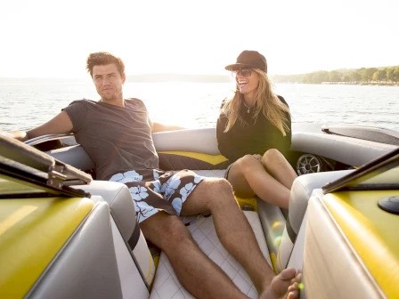 A man and woman are relaxing on a boat, enjoying a sunny day by the water, surrounded by yellow and white upholstery.