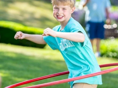 A boy in a blue shirt is joyfully using a hula hoop outside on a sunny day.