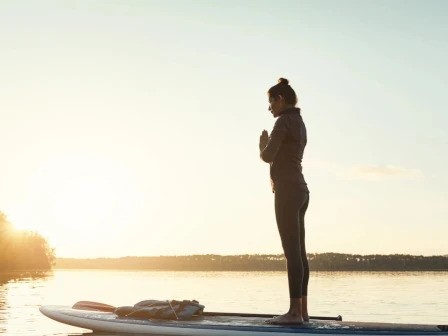 A person stands on a paddleboard in a yoga pose on a calm lake during sunset, hands together in front, creating a serene silhouette.