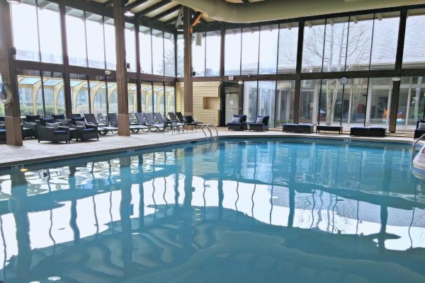 An indoor swimming pool with lounge chairs around it, large windows, and a wooden beam ceiling.