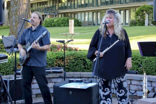 Two musicians perform outdoors: one plays guitar and sings, the other holds a flute. Greenery and a building are in the background.