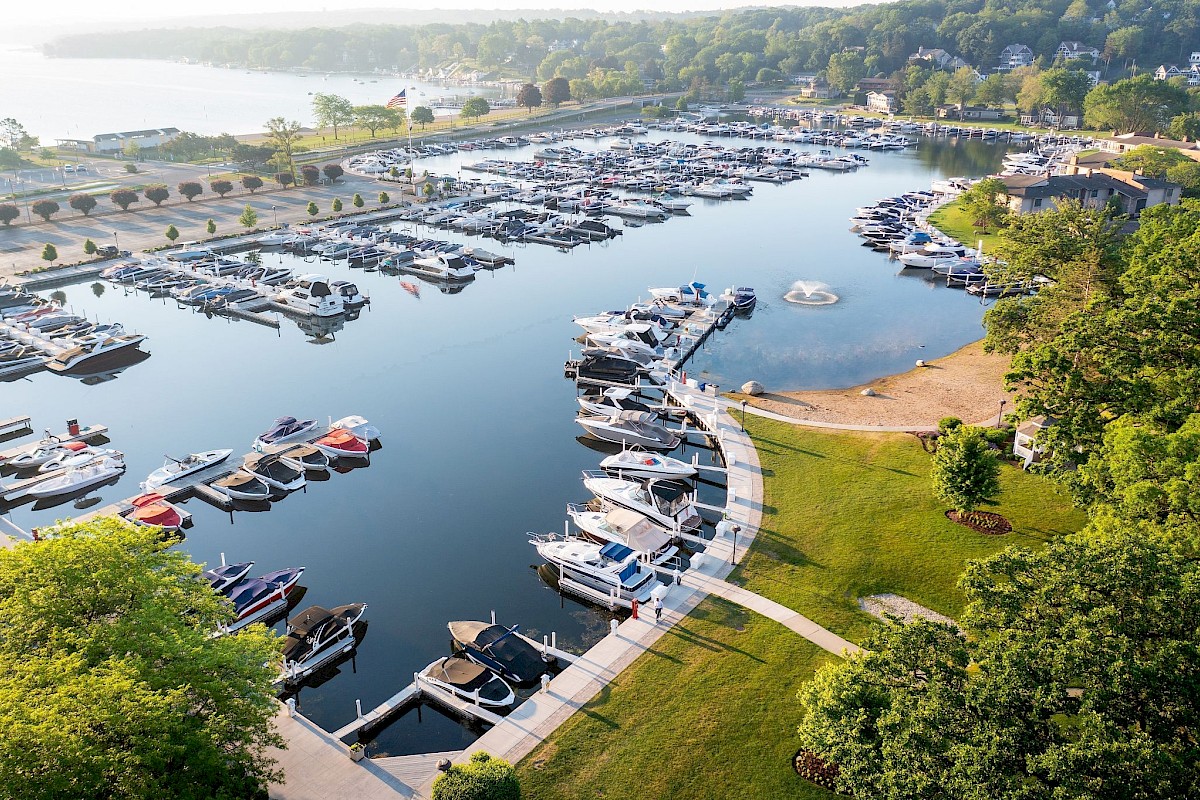 Aerial view of a marina with numerous boats docked in calm water, surrounded by greenery and a park path along the shoreline.
