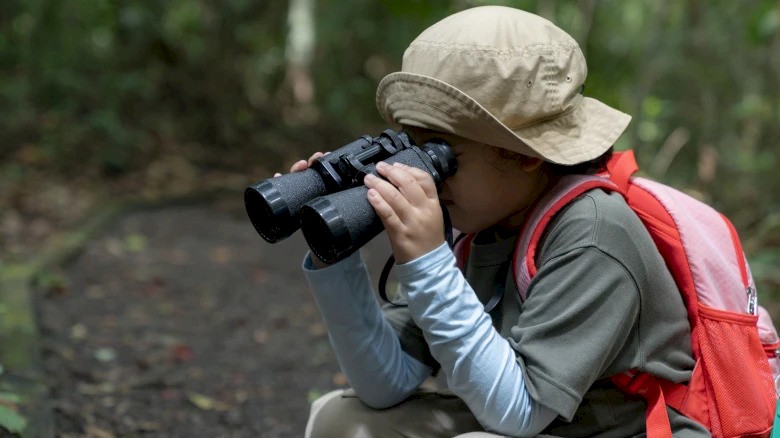 A person with a hat and backpack is using binoculars in a forested area.