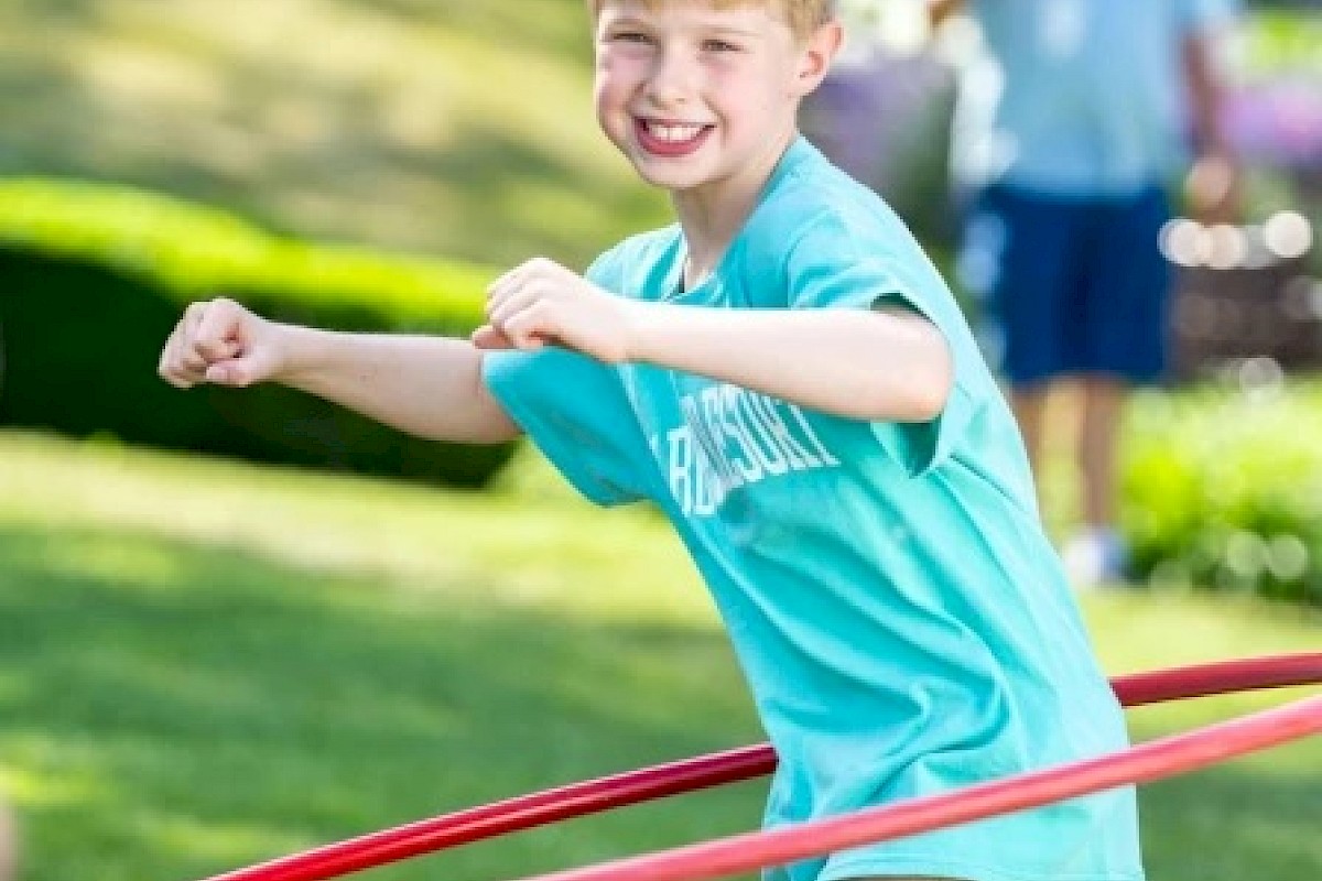 A child happily playing with a hula hoop in a sunny outdoor setting, wearing a blue shirt and smiling broadly at the camera.