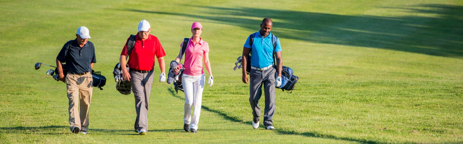 Four golfers are walking together on a golf course, carrying their bags, with a sand trap visible in the background.