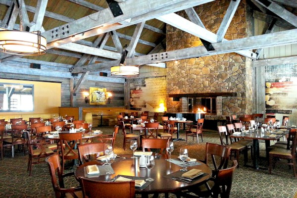The image shows a rustic restaurant interior with wooden beams, stone walls, round tables with settings, and a large fireplace.