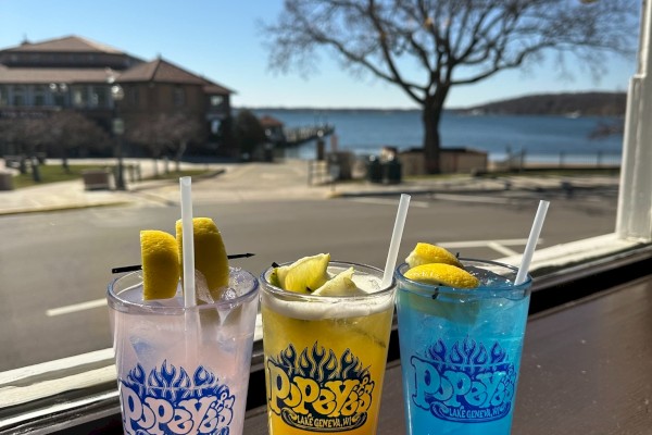 The image shows three colorful drinks in glasses with logos, placed on a ledge with a scenic view of a tree, buildings, and water in the background.
