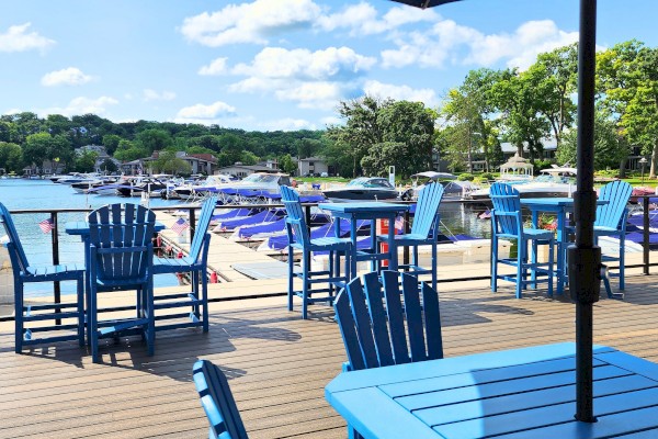This image shows an outdoor seating area with blue tables and chairs overlooking a marina with boats, set against a backdrop of trees and blue skies.